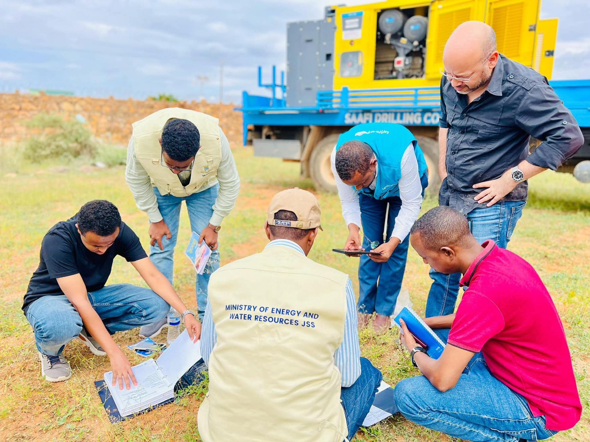 Group of people examining documents and taking notes outdoors near drilling equipment.